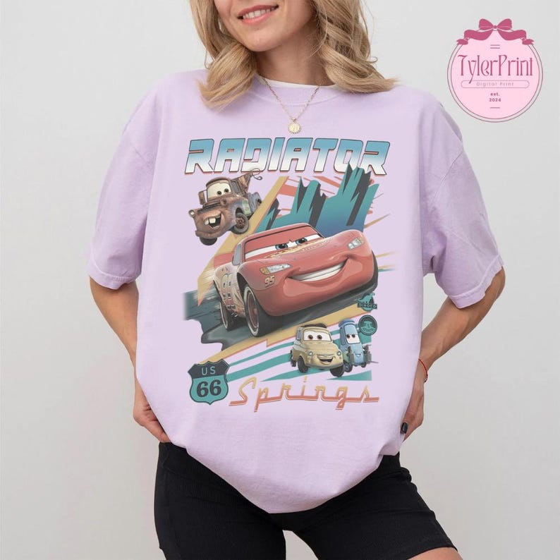 May include: A light purple short-sleeve t-shirt featuring a graphic print of characters from the movie Cars, including Lightning McQueen, Mater, Luigi, and Guido. The design includes the text Radiator Springs and a US 66 road sign emblem.