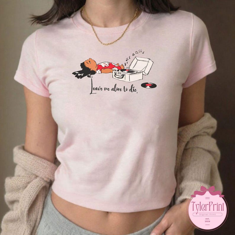 Puede incluir: Camiseta rosa claro con un dibujo de una persona tumbada junto a un tocadiscos. El texto "Leave me alone to die" est&aacute; impreso en la camiseta. Un peque&ntilde;o disco est&aacute; al lado del tocadiscos.