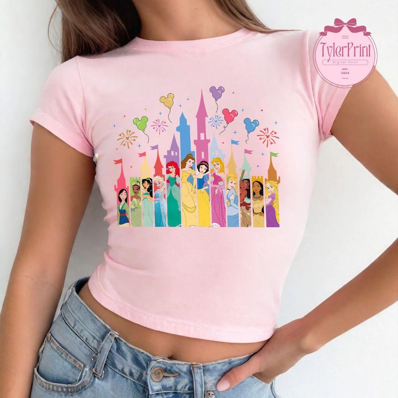 May include: A pink cropped t-shirt featuring a graphic of the Disney castle and all the Disney princesses. The castle is surrounded by fireworks and balloons.