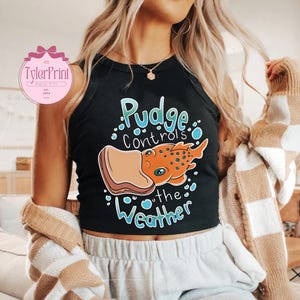 May include: Black crop top featuring the text "Pudge Controls the Weather" and a cartoon illustration of an orange creature with blue eyes and a sandwich. The top has a round neckline and is sleeveless.