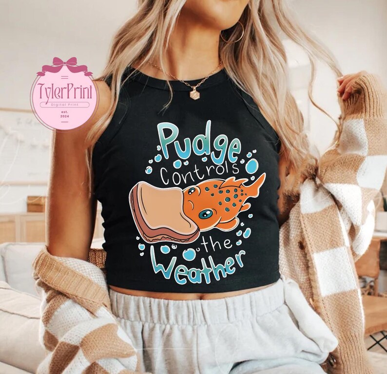 May include: Black crop top featuring the text "Pudge Controls the Weather" and a cartoon illustration of an orange creature with blue eyes and a sandwich. The top has a round neckline and is sleeveless.