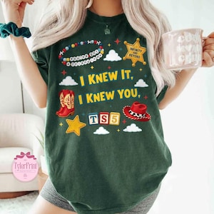 May include: A dark green t-shirt featuring a graphic print with the text "I KNEW IT, I KNEW YOU," "INFINITY AND BEYOND," and "TS5." The design includes a beaded bracelet, a gold sheriff star, red cowboy boots, a red cowboy hat, and blocks.