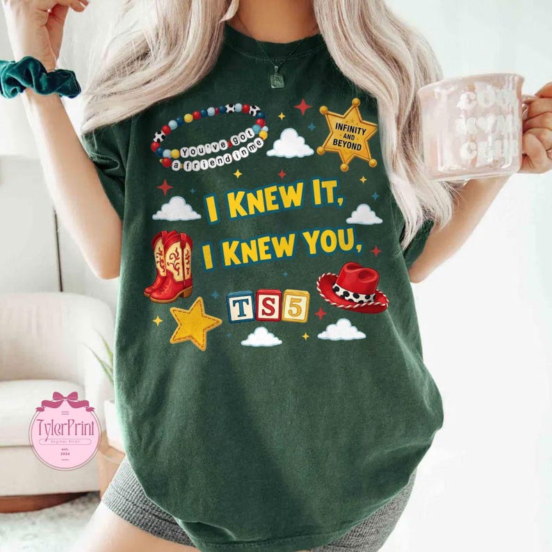 May include: A dark green t-shirt featuring a graphic print with the text "I KNEW IT, I KNEW YOU," "INFINITY AND BEYOND," and "TS5." The design includes a beaded bracelet, a gold sheriff star, red cowboy boots, a red cowboy hat, and blocks.