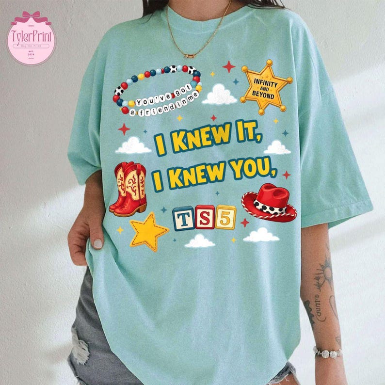 May include: A light blue t-shirt featuring a graphic print with a beaded bracelet, a yellow sheriff star, red cowboy boots, a red cowboy hat, and blocks spelling TS5. The text reads I KNEW IT, I KNEW YOU, and INFINITY AND BEYOND.