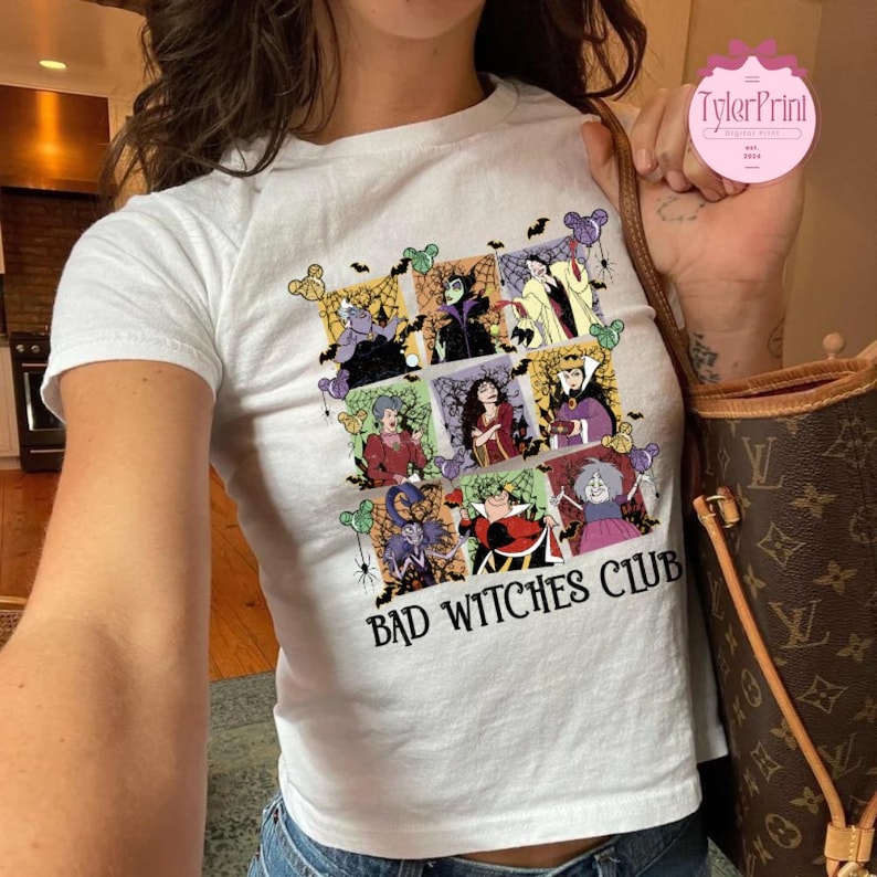 May include: White t-shirt with a colorful graphic featuring various cartoon witches in square frames, with the text "BAD WITCHES CLUB" in a bold font. The shirt has short sleeves.