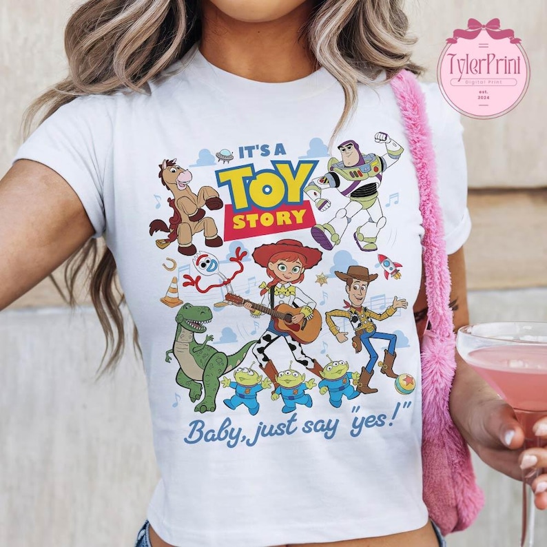 May include: A white short-sleeved t-shirt featuring a colorful graphic print of Toy Story characters including Woody, Jessie, Buzz Lightyear, Rex, Bullseye, and the Aliens. The text reads IT'S A TOY STORY and Baby, just say yes! in blue and red.