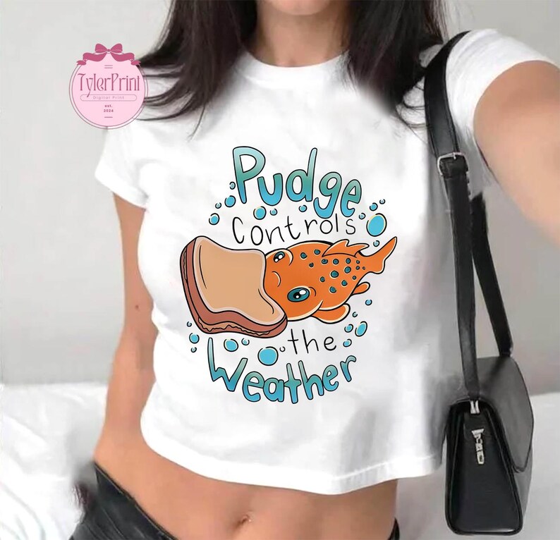 May include: White crop top featuring the text "Pudge Controls the Weather" and an orange fish cartoon with a bread slice. The top has a round neck and short sleeves.