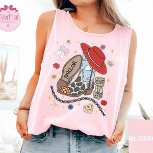May include: A light pink sleeveless tank top featuring a graphic print of a cowboy boot with a cow print pattern, a red hat, a blue sky background, the name Taylor, the number 13 on blocks, and a beaded string with letters spelling out a phrase.