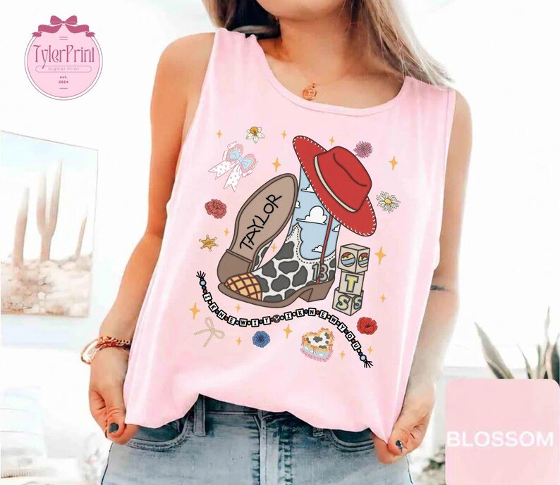 May include: A light pink sleeveless tank top featuring a graphic print of a cowboy boot with a cow print pattern, a red hat, a blue sky background, the name Taylor, the number 13 on blocks, and a beaded string with letters spelling out a phrase.