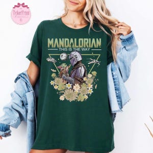 May include: Dark green t-shirt featuring a graphic of The Mandalorian and Grogu surrounded by flowers. The text "MANDALORIAN THIS IS THE WAY" is printed above the image. The shirt has rolled-up sleeves.