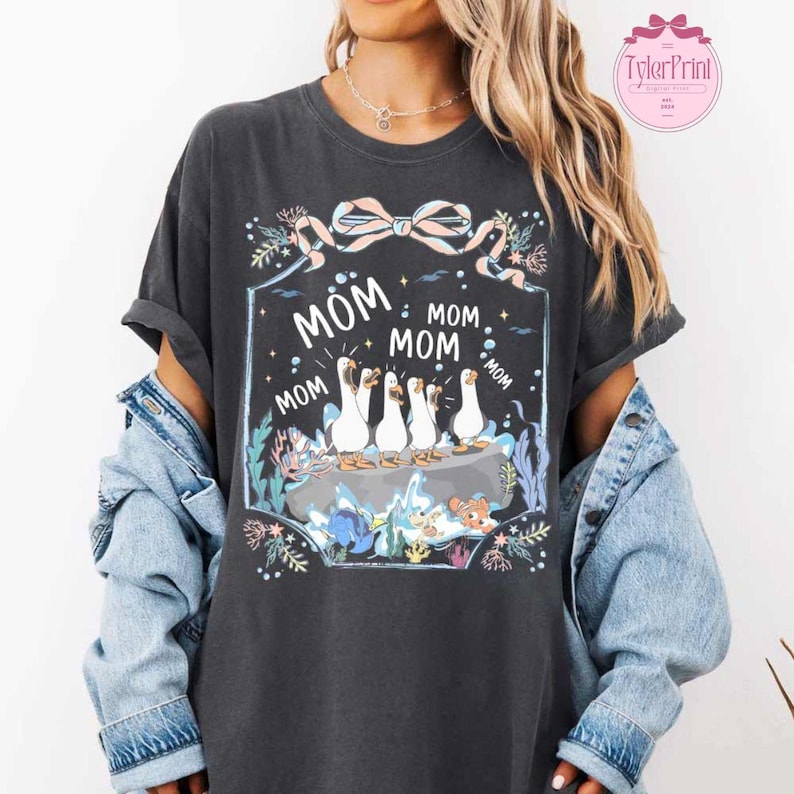 May include: Dark gray t-shirt with a graphic featuring white geese and the word "MOM" repeated. The design includes a decorative bow and floral elements. The shirt is a casual style.