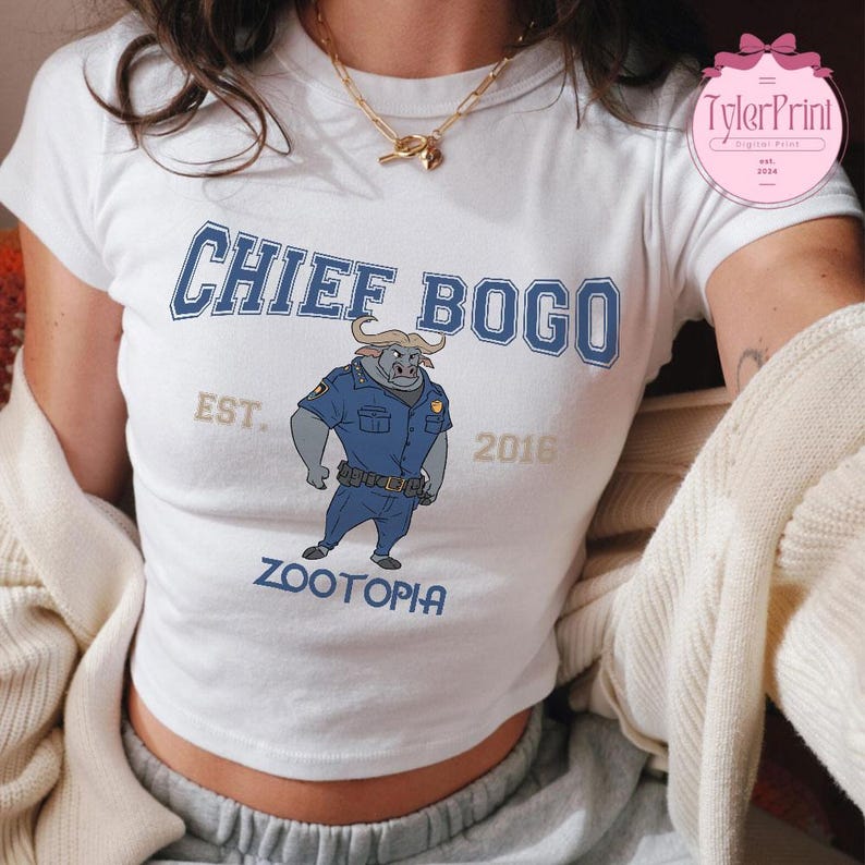 May include: White crop top featuring a blue graphic of Chief Bogo from Zootopia. The text "CHIEF BOGO" is arched across the top, with "EST. 2016" and "ZOOTOPIA" below the illustration. The shirt has short sleeves.