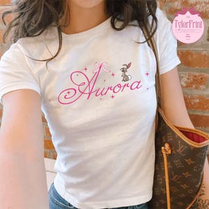 Pu&ograve; includere: Maglietta bianca con un design rosa che presenta il nome "Aurora" in un font corsivo con un piccolo coniglio e un fiocco. Il design include anche piccole stelle rosa.