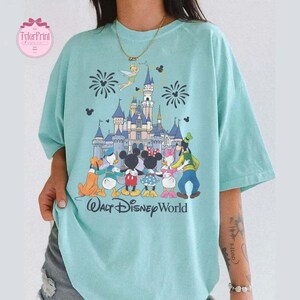 Puede incluir: Camiseta azul claro con un estampado colorido de personajes de Disney frente a un castillo con fuegos artificiales. Las palabras "Walt Disney World" est&aacute;n impresas debajo. La camiseta tiene mangas cortas y un corte holgado.