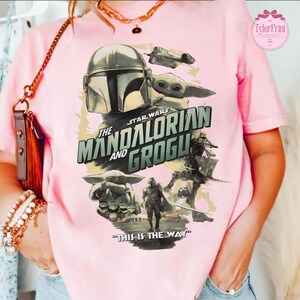 May include: Pink t-shirt featuring a graphic from the Star Wars series, "The Mandalorian and Grogu." The design includes images of the Mandalorian, Grogu, and a spaceship, with the phrase "This is the Way."