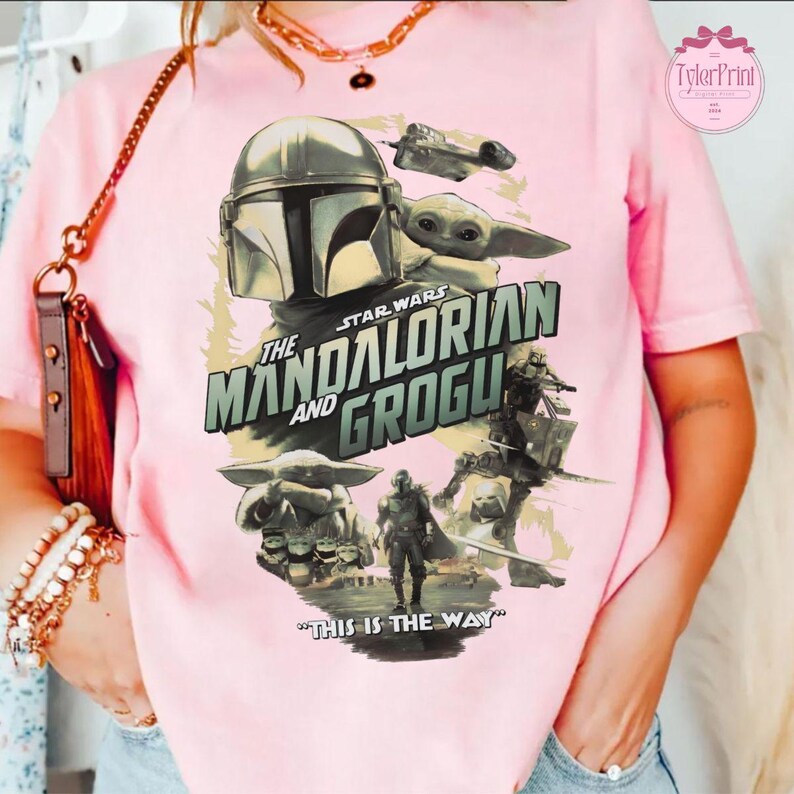 May include: Pink t-shirt featuring a graphic from the Star Wars series, "The Mandalorian and Grogu." The design includes images of the Mandalorian, Grogu, and a spaceship, with the phrase "This is the Way."