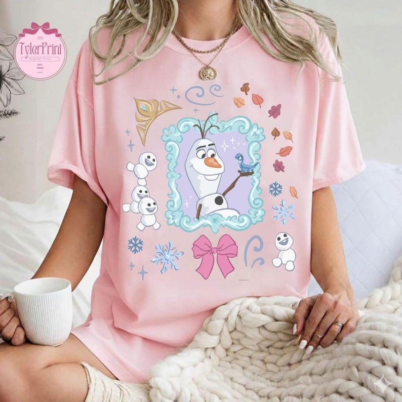 May include: Pink t-shirt featuring a cartoon image of Olaf from Frozen. The design includes Olaf, snowflakes, a blue bird, and a pink bow. The shirt has the text "TylerPrint" in a circular logo.