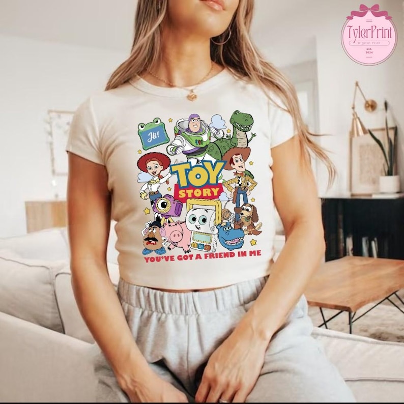 May include: Cream-colored t-shirt featuring a colorful graphic of Toy Story characters. The design includes Woody, Buzz Lightyear, and Rex, with the words "TOY STORY" and "YOU'VE GOT A FRIEND IN ME". The shirt has a crew neck and short sleeves.