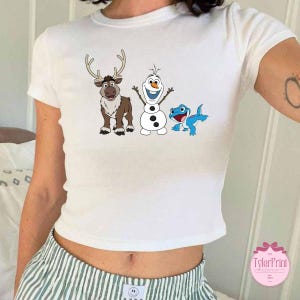 Peut inclure: Haut court blanc avec des personnages de dessins anim&eacute;s du film La Reine des Neiges : Sven le renne, Olaf le bonhomme de neige et une cr&eacute;ature bleue ressemblant &agrave; un l&eacute;zard. Le haut a un col rond et des manches courtes.