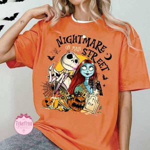 K&ouml;nnte beinhalten: Orangefarbenes T-Shirt mit einem Aufdruck von Jack Skellington und Sally aus The Nightmare Before Christmas, mit dem Text "Nightmare on Main Street". Das Design umfasst K&uuml;rbisse, einen Grabstein und Flederm&auml;use, ideal f&uuml;r Halloween.