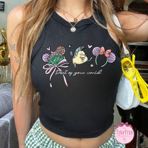 May include: Black tank top with a colorful graphic featuring a mermaid tail, a yellow fish, and mouse ears with a bow. The text "Part of your world" is written in cursive. The tank top is sleeveless.
