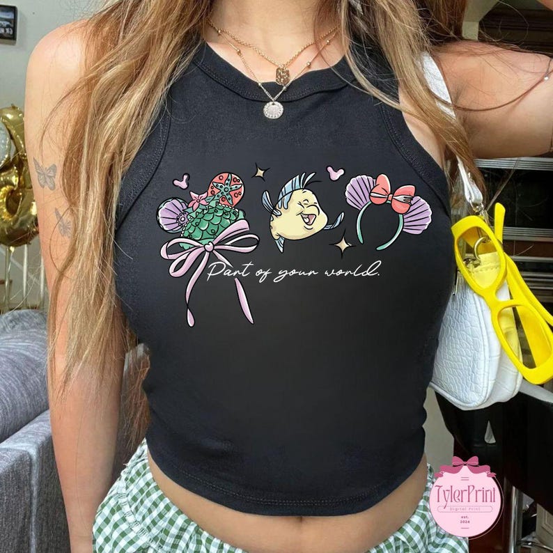 May include: Black tank top with a colorful graphic featuring a mermaid tail, a yellow fish, and mouse ears with a bow. The text "Part of your world" is written in cursive. The tank top is sleeveless.