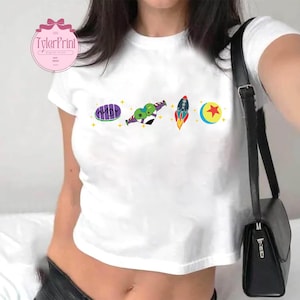 May include: White cropped t-shirt with a graphic print of a cartoon alien, a rocket, a moon, and a star. The text "ANDY" is printed on the shirt.