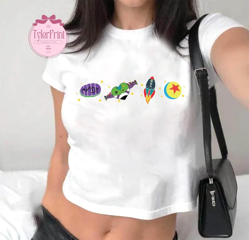 May include: White cropped t-shirt with a graphic print of a cartoon alien, a rocket, a moon, and a star. The text "ANDY" is printed on the shirt.