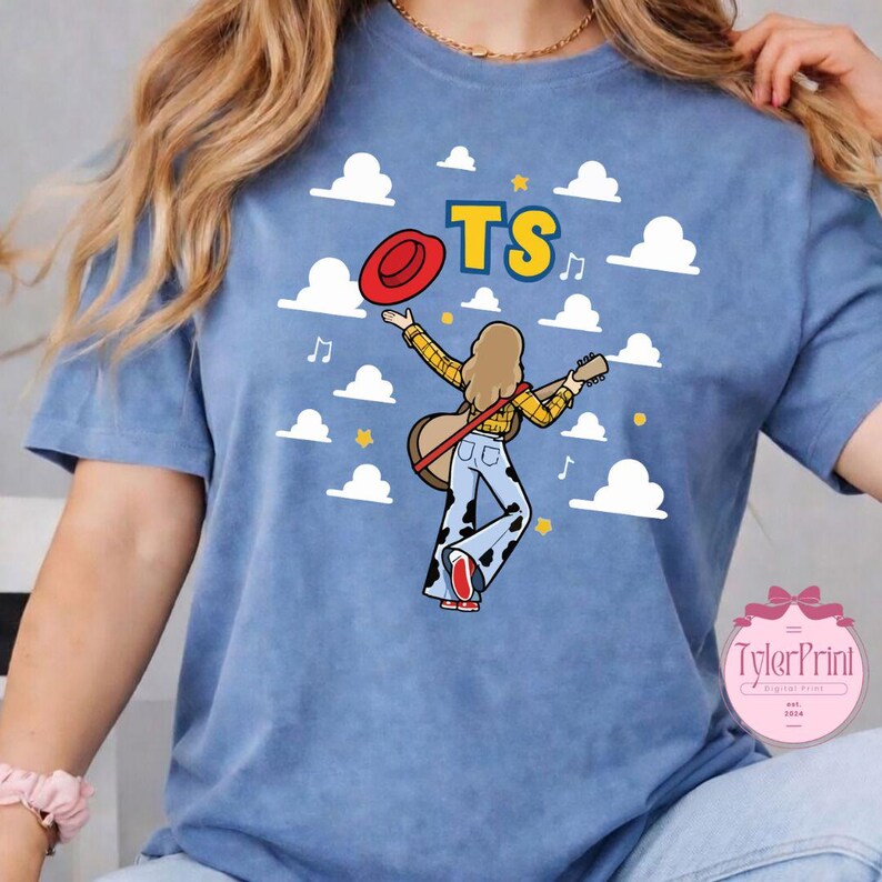 May include: A blue t-shirt featuring a graphic of a person with long hair wearing a yellow plaid shirt and cow-print bell-bottom pants, holding a guitar and a red hat. The design includes white clouds, musical notes, and the yellow text TS.