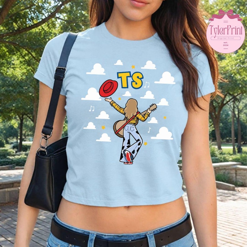 May include: A light blue short-sleeved t-shirt featuring a graphic of a person with long hair wearing a yellow plaid shirt and cow-print pants, holding a guitar and a red hat. The design includes the letters TS and white cloud shapes.