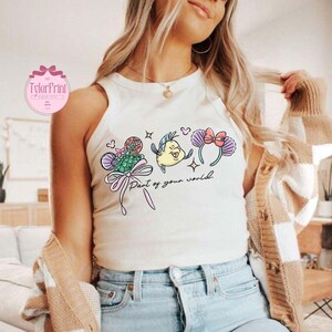 May include: A cream-colored tank top featuring a colorful graphic of cartoon characters and the phrase "Part of your world." The graphic includes a mermaid tail, a fish, and a pair of mouse ears with a bow. The top is worn by a person.
