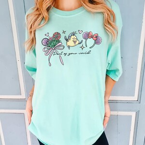 May include: A light teal t-shirt featuring a colorful graphic of cartoon characters and the text "Part of your world." The graphic includes a mermaid tail, a yellow fish, and mouse ears with bows. The shirt has a relaxed fit.
