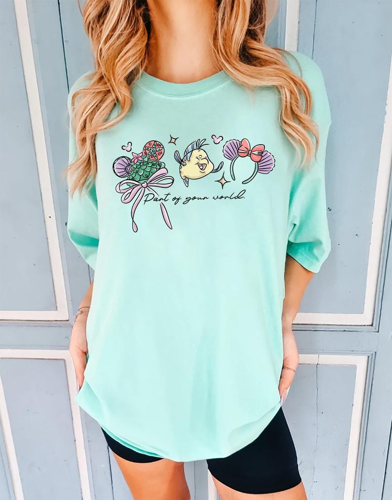 May include: A light teal t-shirt featuring a colorful graphic of cartoon characters and the text "Part of your world." The graphic includes a mermaid tail, a yellow fish, and mouse ears with bows. The shirt has a relaxed fit.