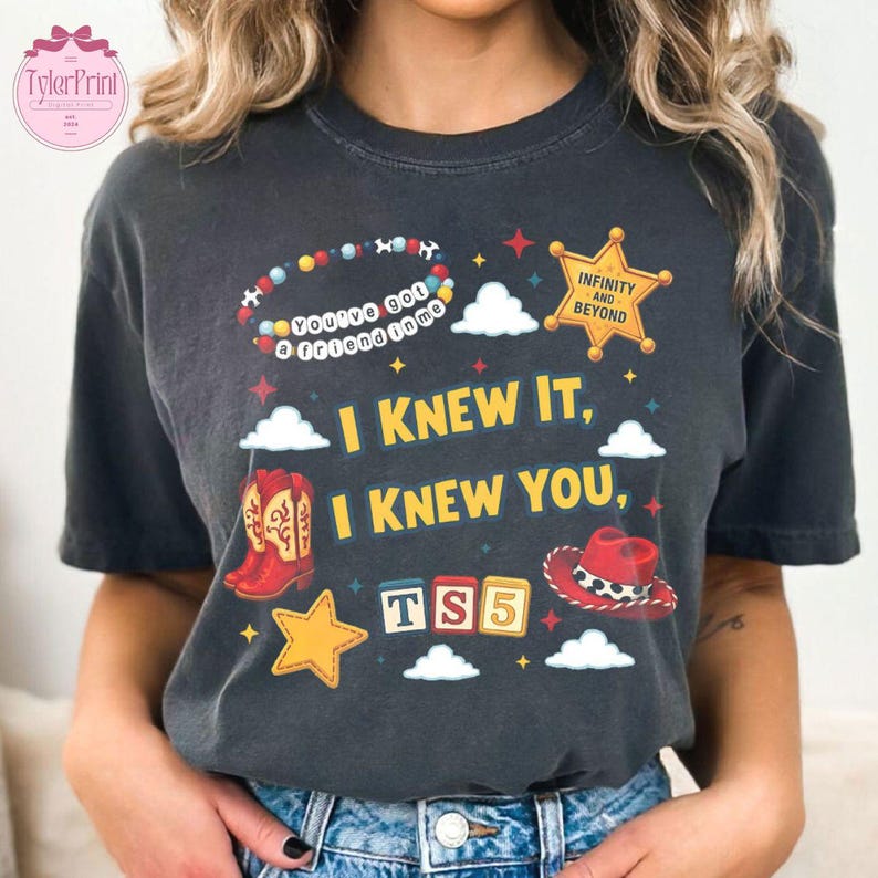 May include: A charcoal grey short-sleeve t-shirt featuring a graphic print with a beaded bracelet reading "You've got a friend in me," a yellow sheriff star badge, red cowboy boots, a red cowboy hat, and the text "I KNEW IT, I KNEW YOU, TS5."