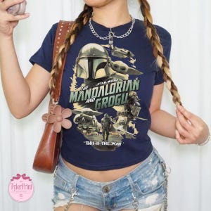May include: Navy blue t-shirt featuring a graphic from the Star Wars series, "The Mandalorian and Grogu." The design includes the Mandalorian's helmet, Grogu, and the phrase "This is the Way."