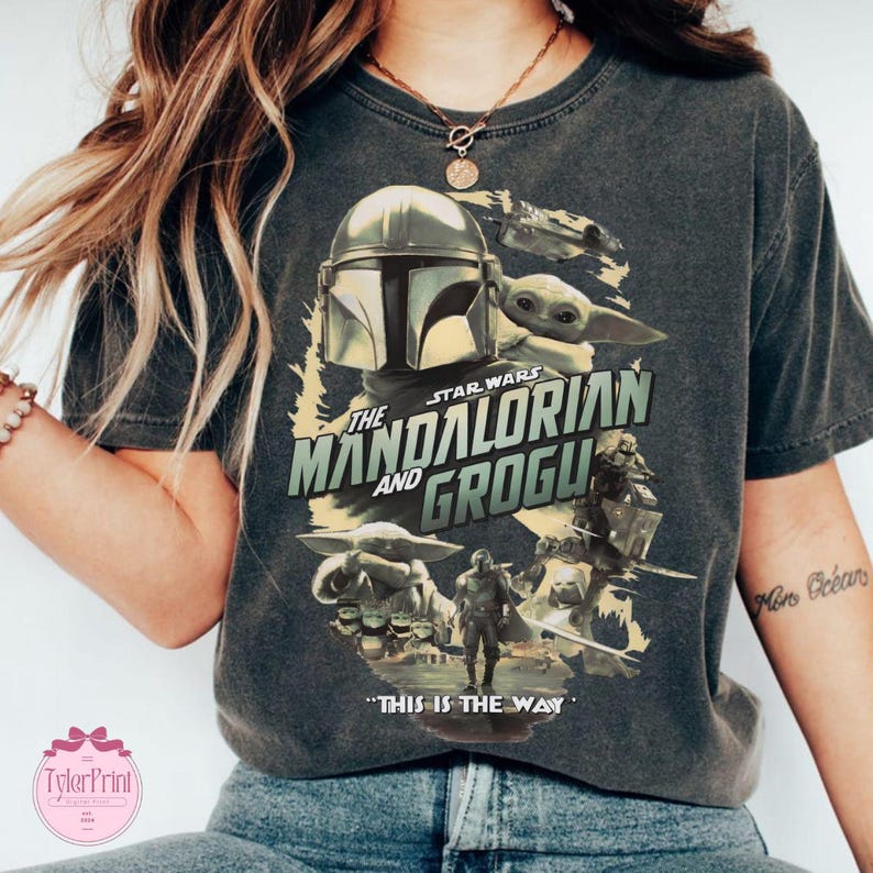 May include: Dark gray t-shirt featuring a graphic from the Star Wars series, "The Mandalorian and Grogu." The design includes the Mandalorian's helmet, Grogu, and the phrase "This is the Way."