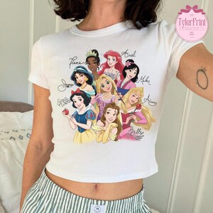May include: White crop top featuring a colorful graphic of Disney princesses, including Snow White, Belle, and Ariel. The names of the princesses are written in cursive. The shirt has a round neckline and short sleeves.