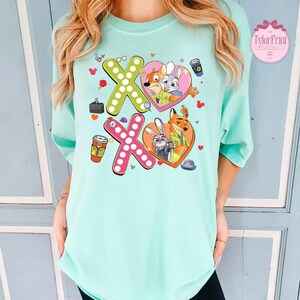 May include: A light teal t-shirt featuring a colorful graphic design. The design includes the letters "XO" in green and pink, with cartoon characters inside heart shapes. Additional elements include coffee cups and a small briefcase.