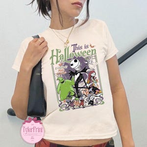 K&ouml;nnte beinhalten: Cremefarbenes T-Shirt mit einem Grafikdesign aus "The Nightmare Before Christmas". Das Design zeigt Jack Skellington, Sally und andere Charaktere mit dem Text "This is Halloween". Das Shirt hat kurze &Auml;rmel.