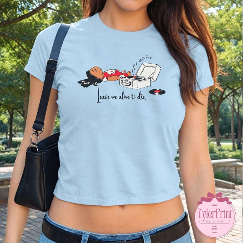 Puede incluir: Camiseta azul claro con un gr&aacute;fico de una persona acostada junto a un tocadiscos y un disco. El texto "Leave me alone to die" est&aacute; impreso en la camiseta.