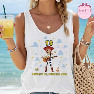 May include: A white sleeveless tank top featuring a graphic of a cowgirl character playing an acoustic guitar. The design includes the text TS and I Knew It, I Knew You, surrounded by blue cloud shapes, musical notes, a ball, and a traffic cone.