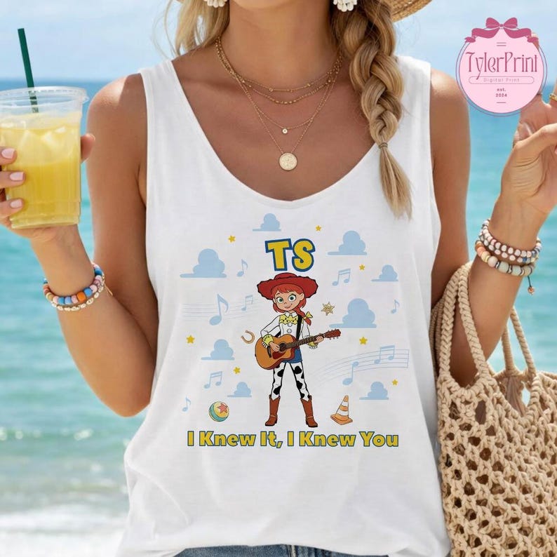 May include: A white sleeveless tank top featuring a graphic of a cowgirl character playing an acoustic guitar. The design includes the text TS and I Knew It, I Knew You, surrounded by blue cloud shapes, musical notes, a ball, and a traffic cone.