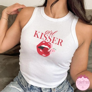 Peut inclure: Débardeur blanc avec l'inscription rouge "girl KISSER" au-dessus d'une image de lèvres rouges et d'une cerise avec un nœud rose. Le débardeur est sans manches et a une encolure ronde.