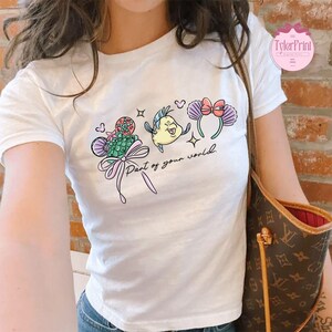 May include: White t-shirt featuring a colorful graphic of cartoon characters and the text "Part of your world." The design includes a fish, a seashell, and a headband with a bow. The shirt is a crew neck.