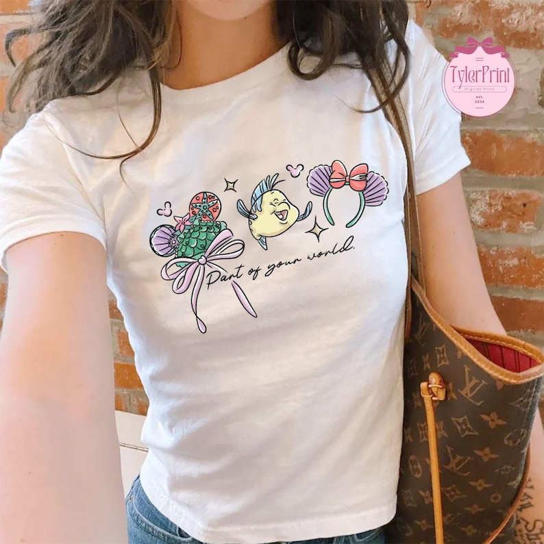May include: White t-shirt featuring a colorful graphic of cartoon characters and the text "Part of your world." The design includes a fish, a seashell, and a headband with a bow. The shirt is a crew neck.