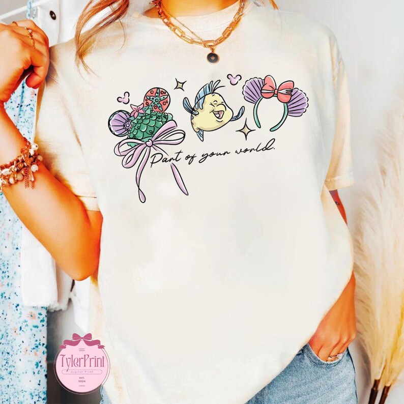 May include: Cream-colored t-shirt featuring a colorful graphic of characters from a popular animated film. The design includes a mermaid, a yellow fish, and mouse ears with a bow, with the text "Part of your world."