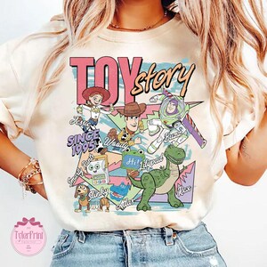 K&ouml;nnte beinhalten: Ein cremefarbenes T-Shirt mit einem bunten Toy Story Grafikdruck. Das Design zeigt die Figuren Jessie, Woody, Buzz Lightyear, Slinky Dog, Rex und andere mit dem Text Toy Story, Since 1995, Jessie, Woody, Buzz, Slinky, Rex und Hi!