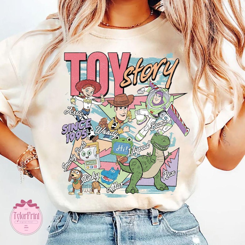 K&ouml;nnte beinhalten: Ein cremefarbenes T-Shirt mit einem bunten Toy Story Grafikdruck. Das Design zeigt die Figuren Jessie, Woody, Buzz Lightyear, Slinky Dog, Rex und andere mit dem Text Toy Story, Since 1995, Jessie, Woody, Buzz, Slinky, Rex und Hi!