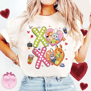 May include: Cream-colored t-shirt featuring a colorful graphic of characters from Zootopia, with the letters "XO" and heart shapes. The design includes cartoon images of Nick Wilde, Judy Hopps, and other themed elements.