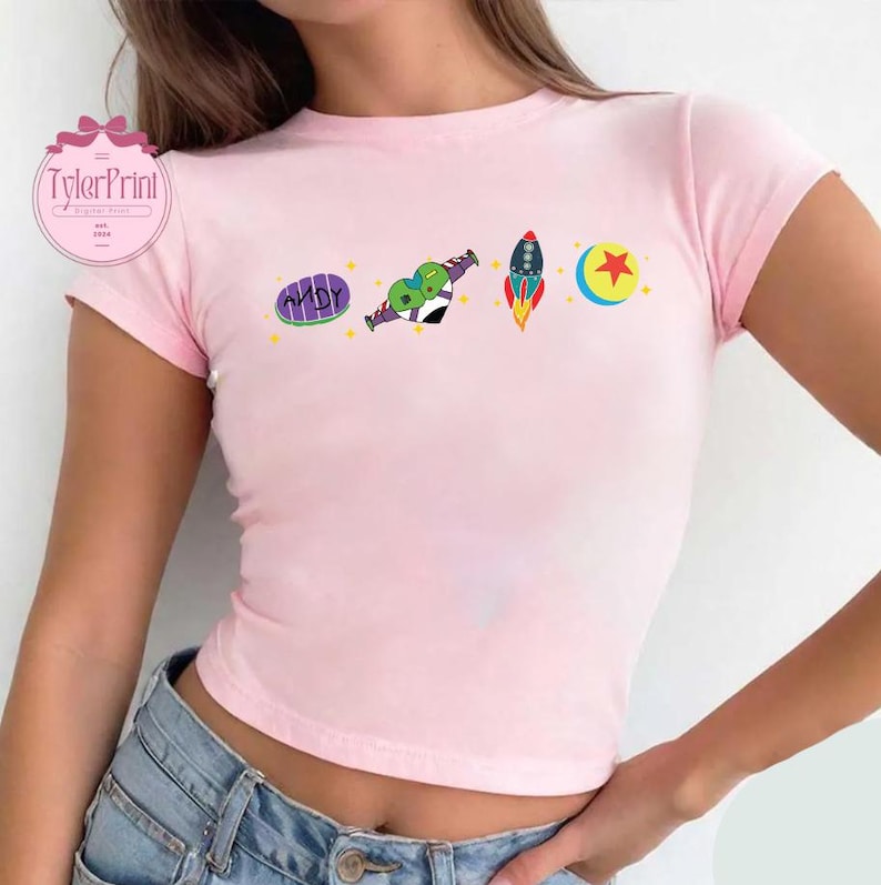 May include: Pink cropped t-shirt with a graphic print of characters from the movie Toy Story. The graphic includes a rocket, a star, a ball, and the words "ANDY" on a pizza.